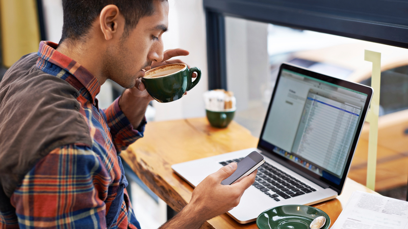 Seated man sipping coffee in front of window with a laptop, using a smart phone.